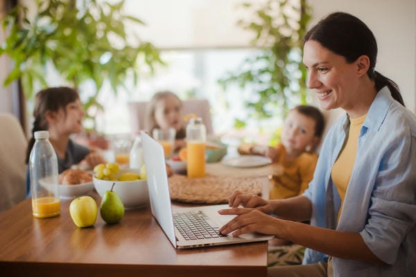 arbeitnehmer-elternzeit Eine Mutter sitz am Küchentisch mit ihren drei Kindern während der Elternzeit und plant die Rückkehr ins Berufsleben an ihrem Laptop.