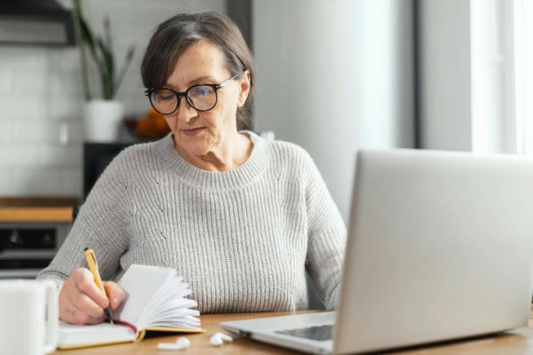 arbeitnehmer-generation-50plus Eine Frau der Generation 50plus mit Brille sitzt an einem Tisch vor einem Laptop und schreibt konzentriert in ein Notizbuch.