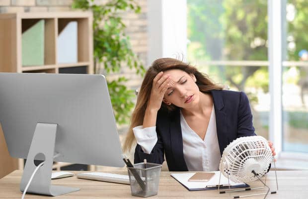 arbeitsplatz-hitze-im-buero Eine erschöpfte Frau sitzt wegen der Hitze am Büroschreibtisch und richtet einen kleinen Tischventilator auf ihr Gesicht.