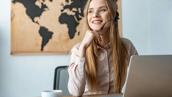 Eine Tourismuskauffrau mit Headset vorm Laptop. Im Hintergrund ist eine Weltkarte zu sehen.