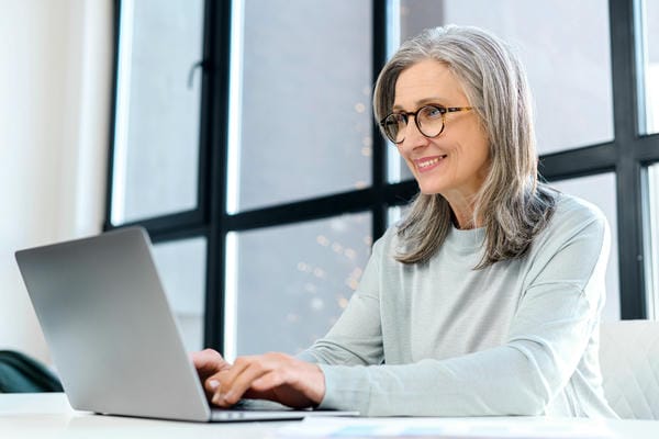 bewerbung-50plus Eine lächelnde, ältere Frau mit grauen Haaren und Brille schreibt konzentriert ein Bewerbung 50plus an einem Laptop.