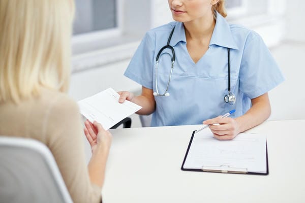 gesundheit-arbeitsunfaehigkeit Eine medizinische Fachkraft in blauer Arbeitskleidung übergibt einer Patientin eine Bescheinigung über deren Arbeitsunfähigkeit.