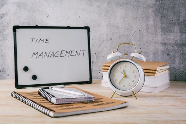 kompetenzen-zeitmanagement Ein weißer Wecker, Notizhefte und ein Whiteboard mit der Aufschrift „TIME MANAGEMENT“ - Zeitmanagement - stehen auf einem hellen Holztisch vor einer grauen Betonwand.